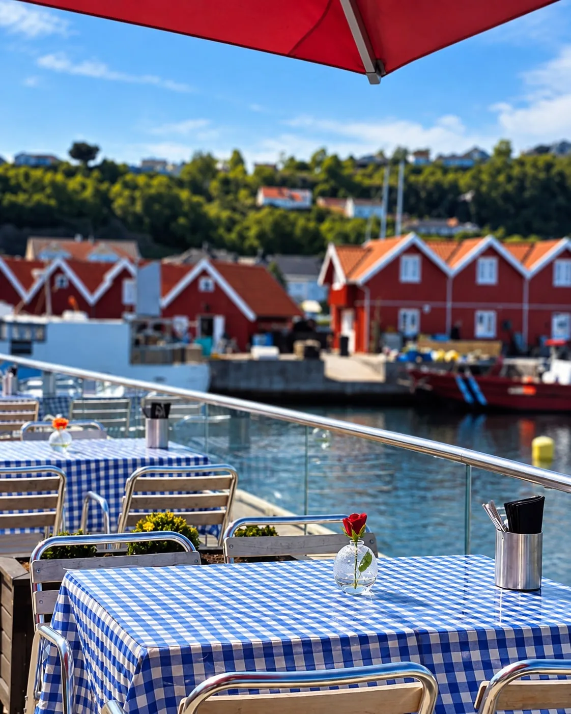 Terrassen til Belmare ved vannet, med blåhvite rutete duker og røde naust på den andre siden.